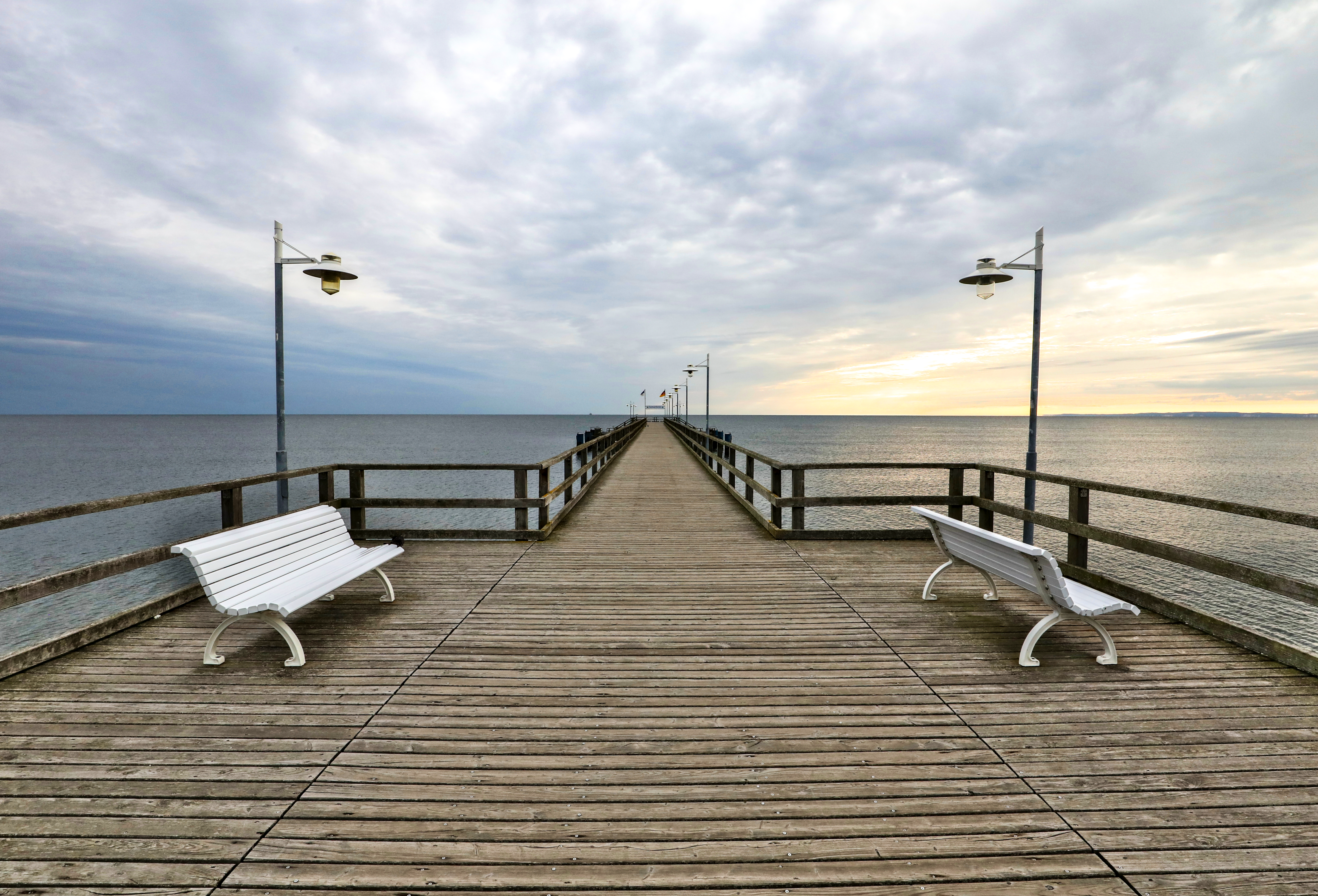 Das Bild wurde auf der Seebrücke stehend aufgenommen und zeigt die gesamte Länge der Brücke. Links und rechts stehen zwei weiße Bänke, die Sonne geht langsam unter und die Brücke ist umgeben von der Ostsee